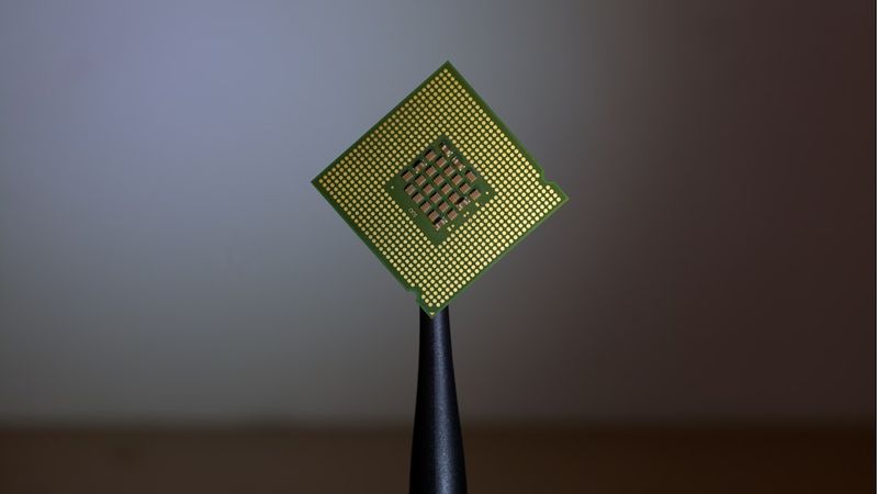 The image features a close-up of a computer processor being held by tweezers against a soft, blurred background. The processor is a small, rectangular chip with a grid of gold pins visible on its underside.