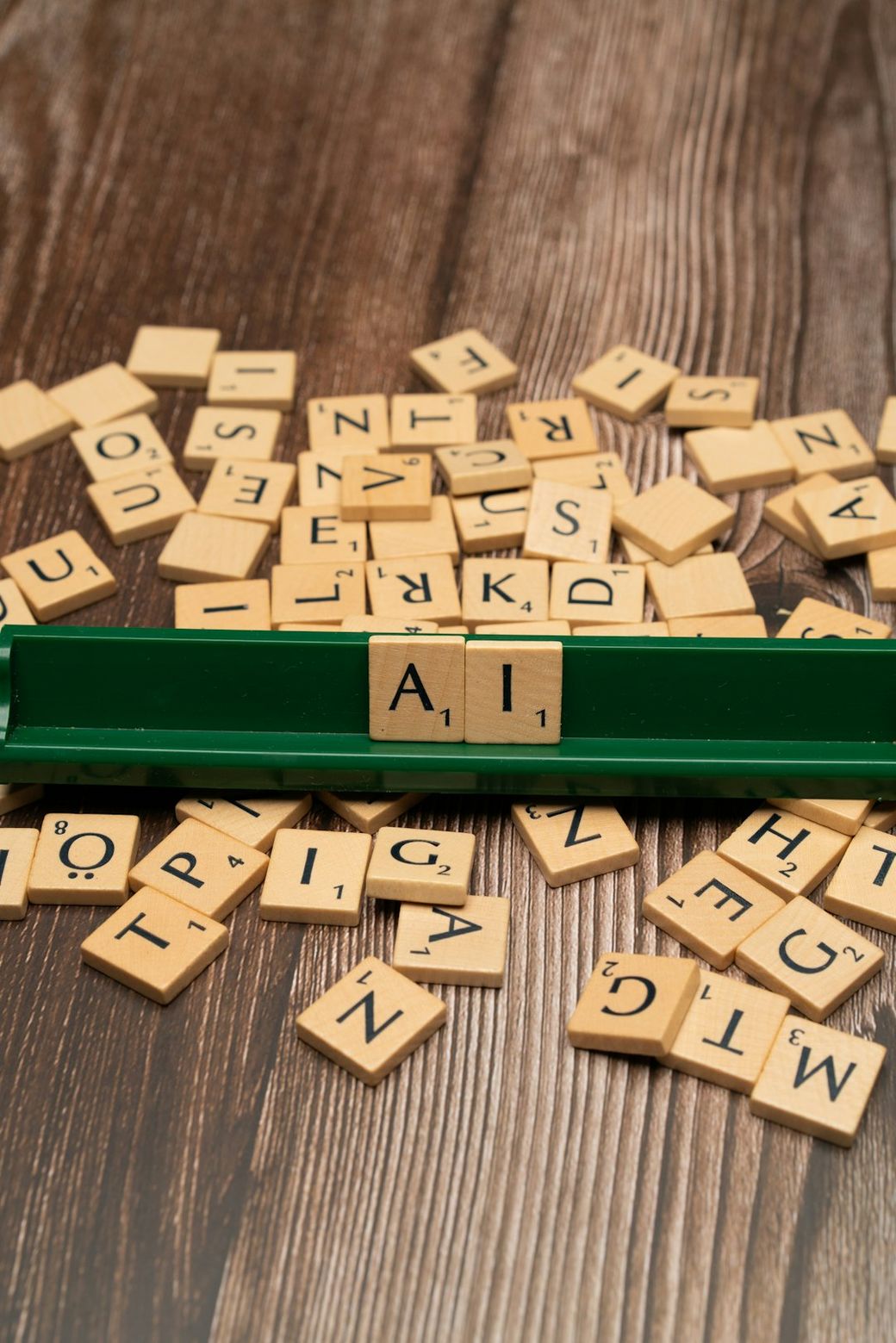 The image features a collection of wooden letter tiles scattered on a wooden surface, with a green tile holder displaying the letters 'A', 'I'. The arrangement suggests a game of word formation, likely Scrabble. — Credit: Photo by Markus Winkler on Unsplash