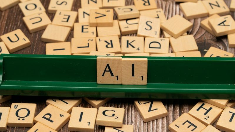 The image features a collection of wooden letter tiles scattered on a wooden surface, with a green tile holder displaying the letters 'A', 'I'. The arrangement suggests a game of word formation, likely Scrabble.