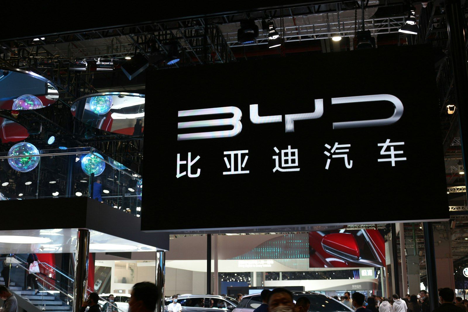 The image showcases a prominent display booth from BYD, a Chinese automotive manufacturer, at an auto show. The booth features a large illuminated sign with the BYD logo and Chinese characters, drawing attention from attendees.
