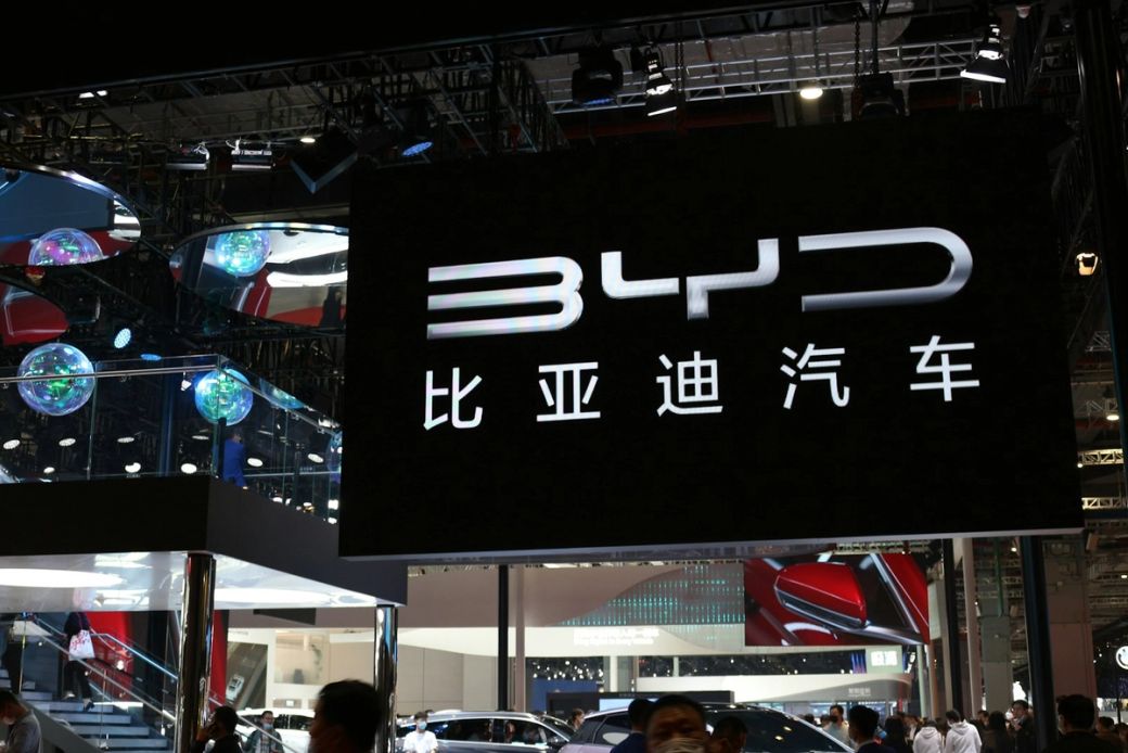 The image showcases a prominent display booth from BYD, a Chinese automotive manufacturer, at an auto show. The booth features a large illuminated sign with the BYD logo and Chinese characters, drawing attention from attendees. — Credit: Photo by P. L. / Unsplash