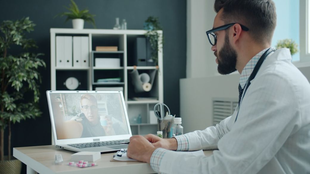 A healthcare professional is shown sitting at a desk in an office setting, engaging with a patient via video call on a laptop. The scene includes elements of a professional workspace, such as medical supplies and plants. — Credit: Photo by Vitaly Gariev on Unsplash