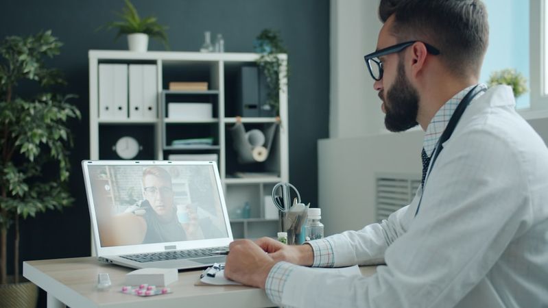 A healthcare professional is shown sitting at a desk in an office setting, engaging with a patient via video call on a laptop. The scene includes elements of a professional workspace, such as medical supplies and plants.