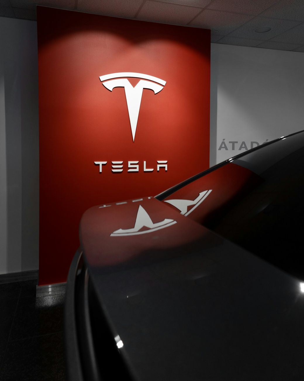 The image features a Tesla logo displayed prominently on a red wall, with a sleek vehicle in the foreground partially obscured. The modern setting suggests a showroom or promotional display. — Credit: Photo by Milan Csizmadia / Unsplash