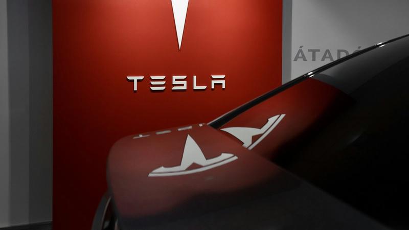 The image features a Tesla logo displayed prominently on a red wall, with a sleek vehicle in the foreground partially obscured. The modern setting suggests a showroom or promotional display.