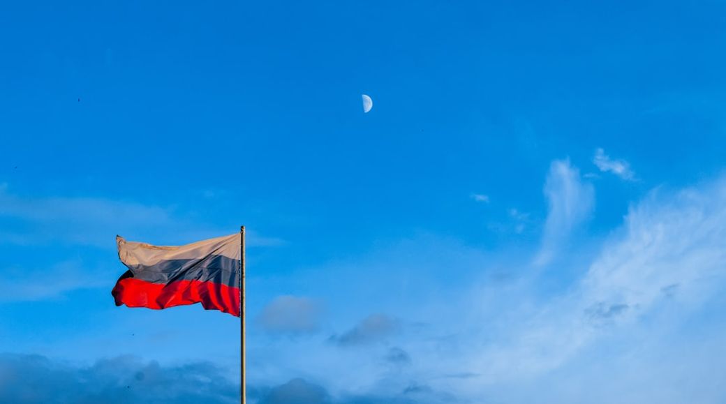 The image features the flag of Russia waving against a clear blue sky with a visible crescent moon. The flag is prominently displayed on a flagpole, symbolizing national pride. — Credit: Photo by Egor Filin on Unsplash