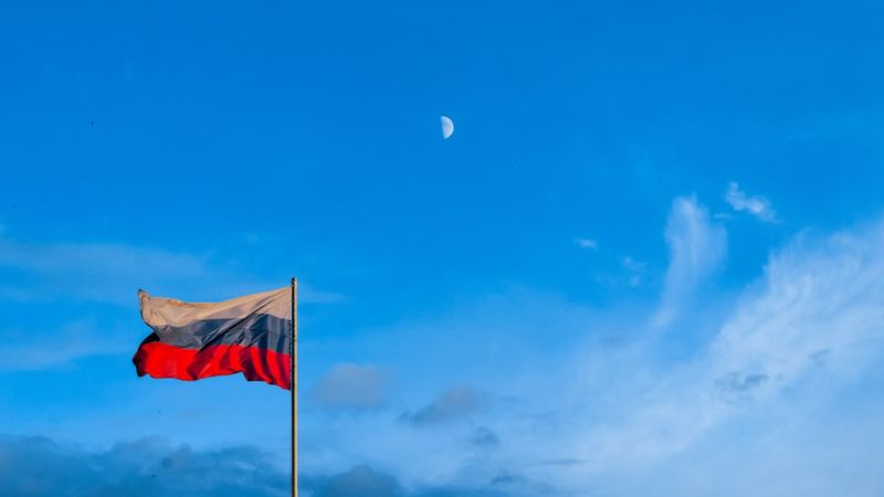 The image features the flag of Russia waving against a clear blue sky with a visible crescent moon. The flag is prominently displayed on a flagpole, symbolizing national pride.