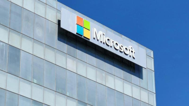 The image shows the exterior of a Microsoft building with a clear blue sky in the background. The prominent Microsoft logo, featuring four colored squares, is displayed on the facade.