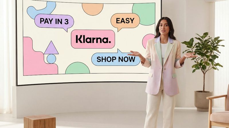 A woman stands in a modern interior space, gesturing towards a large screen displaying colorful shapes and promotional text. The screen features the Klarna logo alongside phrases such as 'PAY IN 3' and 'EASY', indicating a shopping or payment service.