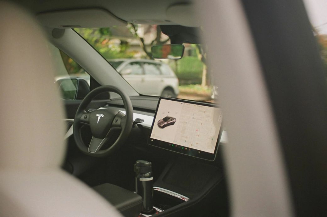 The image shows the interior of a Tesla vehicle, featuring the steering wheel and a large touchscreen display. The screen displays a map and a vehicle model, illustrating the car's digital interface. — Credit: Photo by Murphy Zheng / Unsplash
