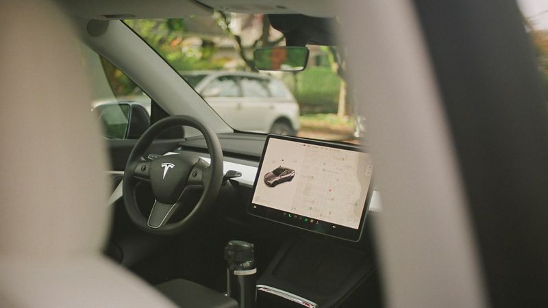 The image shows the interior of a Tesla vehicle, featuring the steering wheel and a large touchscreen display. The screen displays a map and a vehicle model, illustrating the car's digital interface.