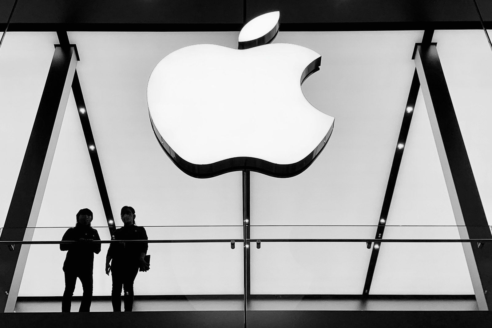 The image depicts two individuals standing on a balcony overlooking an Apple retail store entrance, highlighted by a large illuminated Apple logo. The monochromatic color scheme emphasizes the modern architectural design of the store.