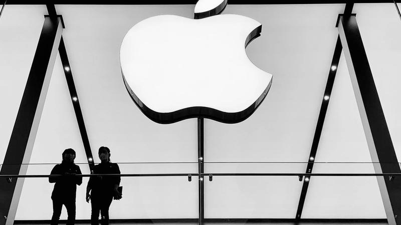 Two silhouettes of people stand on a balcony in front of a large illuminated Apple logo, showcasing the iconic design of an Apple store. The image is in black and white, enhancing the minimalist aesthetic.