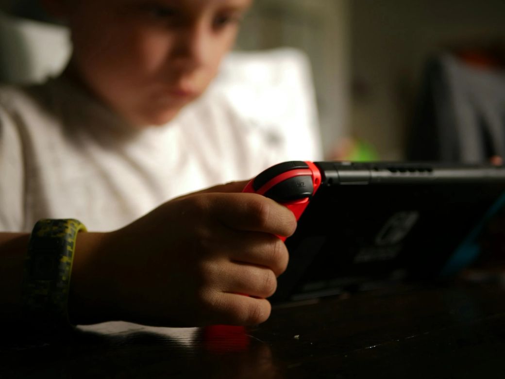 A child is focused on playing a handheld gaming console, holding the device with one hand while using the controls. The background is softly blurred, emphasizing the child's engagement with the screen. — Credit: Photo by Tyler Lagalo / Unsplash