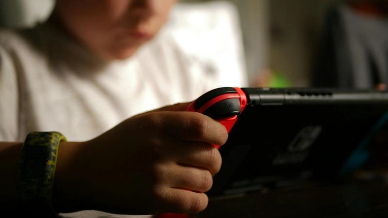 A child is focused on playing a handheld gaming console, holding the device with one hand while using the controls. The background is softly blurred, emphasizing the child's engagement with the screen.