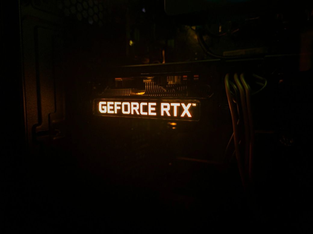 The image features a close-up view of a computer graphics card, specifically highlighting the illuminated 'GEFORCE RTX' branding. The lighting emphasizes the technology and cooling components within the system. — Credit: Photo by Alex Konokh / Unsplash