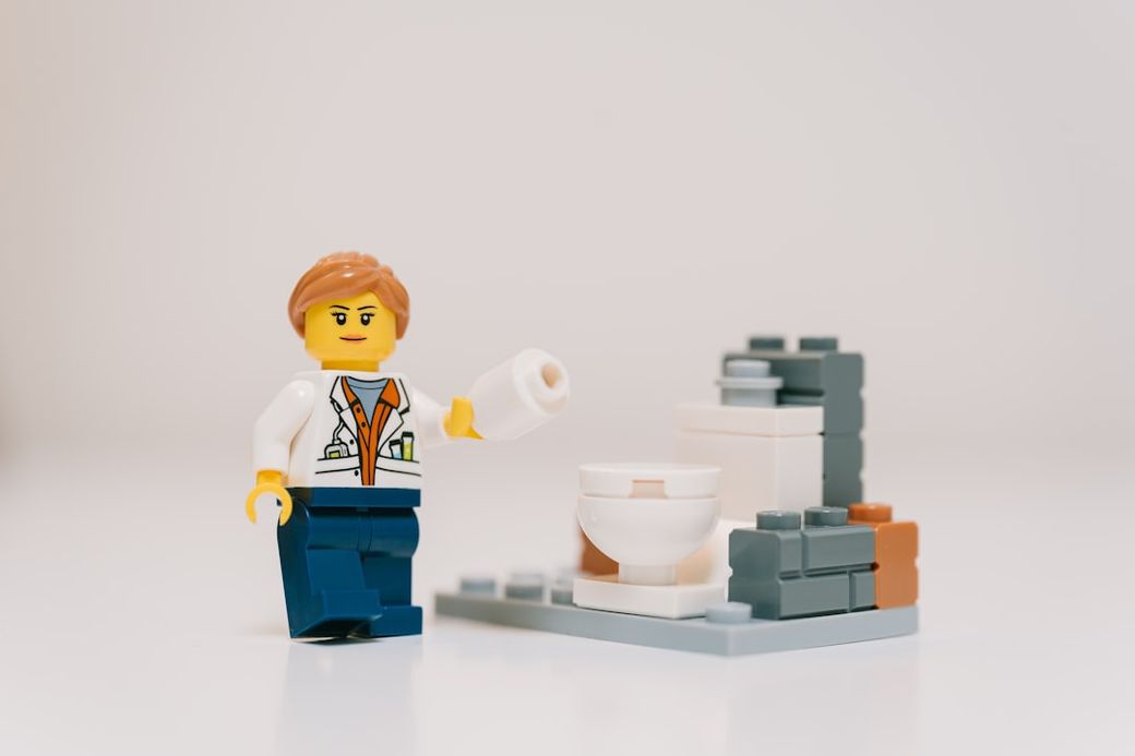 The image features a LEGO figure holding a roll of toilet paper next to a small model of a toilet. The figure appears to be engaging with the bathroom setting in a playful manner. — Credit: Photo by Daniel Cheung on Unsplash