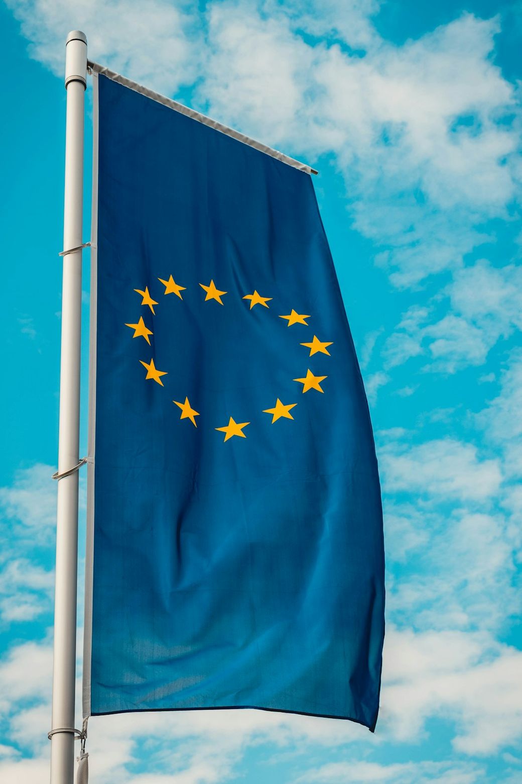 The image features a flag of the European Union waving against a blue sky with scattered clouds. The flag is prominently displayed and features a circle of twelve yellow stars on a blue background. — Credit: Photo by Christian Wiediger on Unsplash