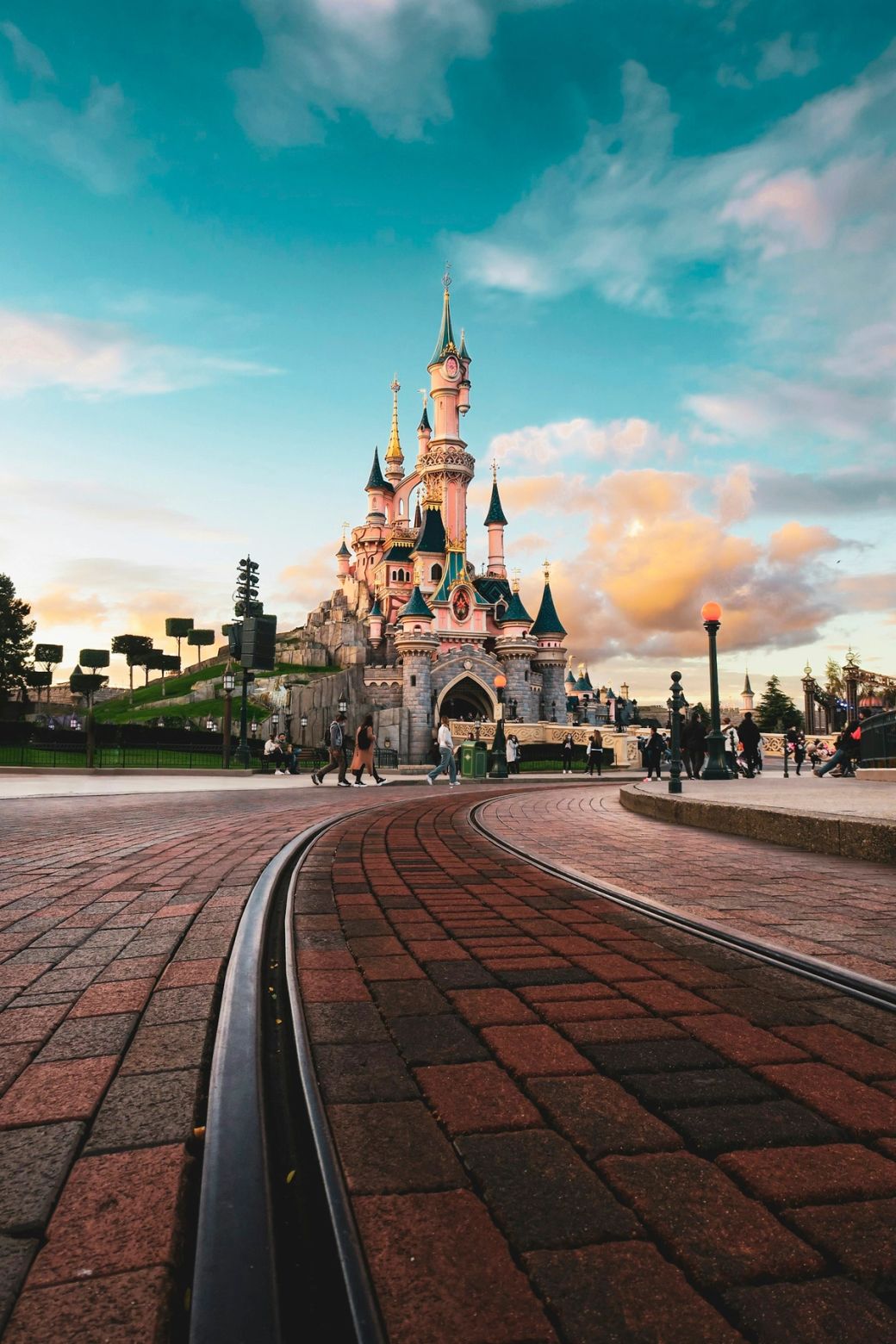 A picturesque view of a fairy-tale castle at a theme park, with visitors strolling in the foreground. The scene captures the vibrant colors of the sky during sunset, enhancing the enchanting atmosphere surrounding the landmark. — Credit: Photo by Bastien Nvs / Unsplash