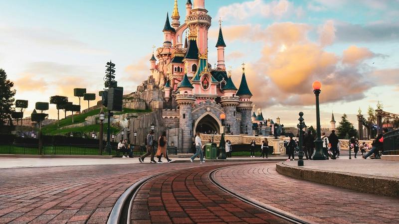 A picturesque view of a fairy-tale castle at a theme park, with visitors strolling in the foreground. The scene captures the vibrant colors of the sky during sunset, enhancing the enchanting atmosphere surrounding the landmark.