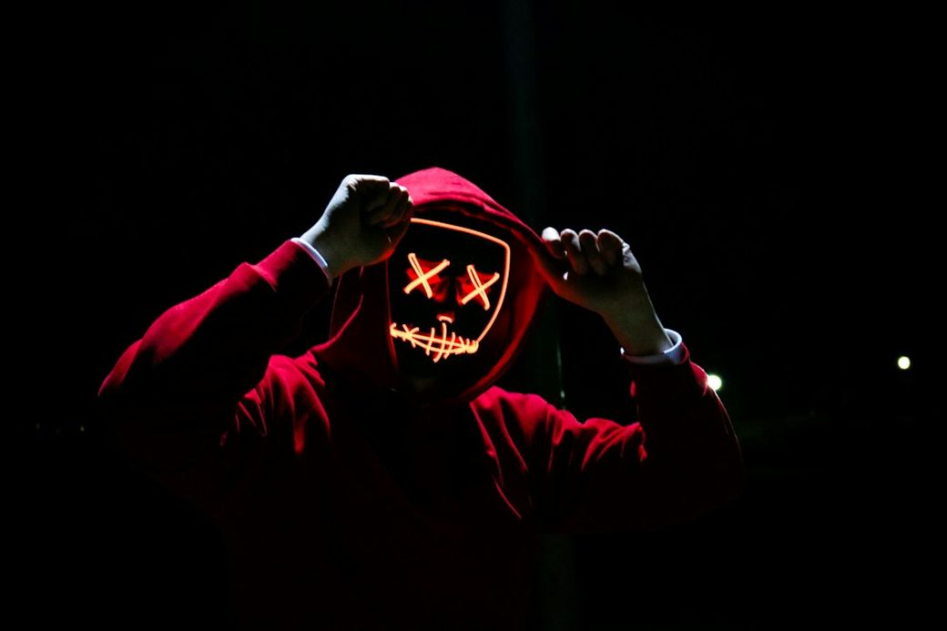 A person wearing a red hoodie is standing against a dark background, holding the hood up while displaying an illuminated mask with a mischievous grin and crossed-out eyes. The scene is set at night with faint lights in the background. — Credit: Photo by sebastiaan stam on Unsplash
