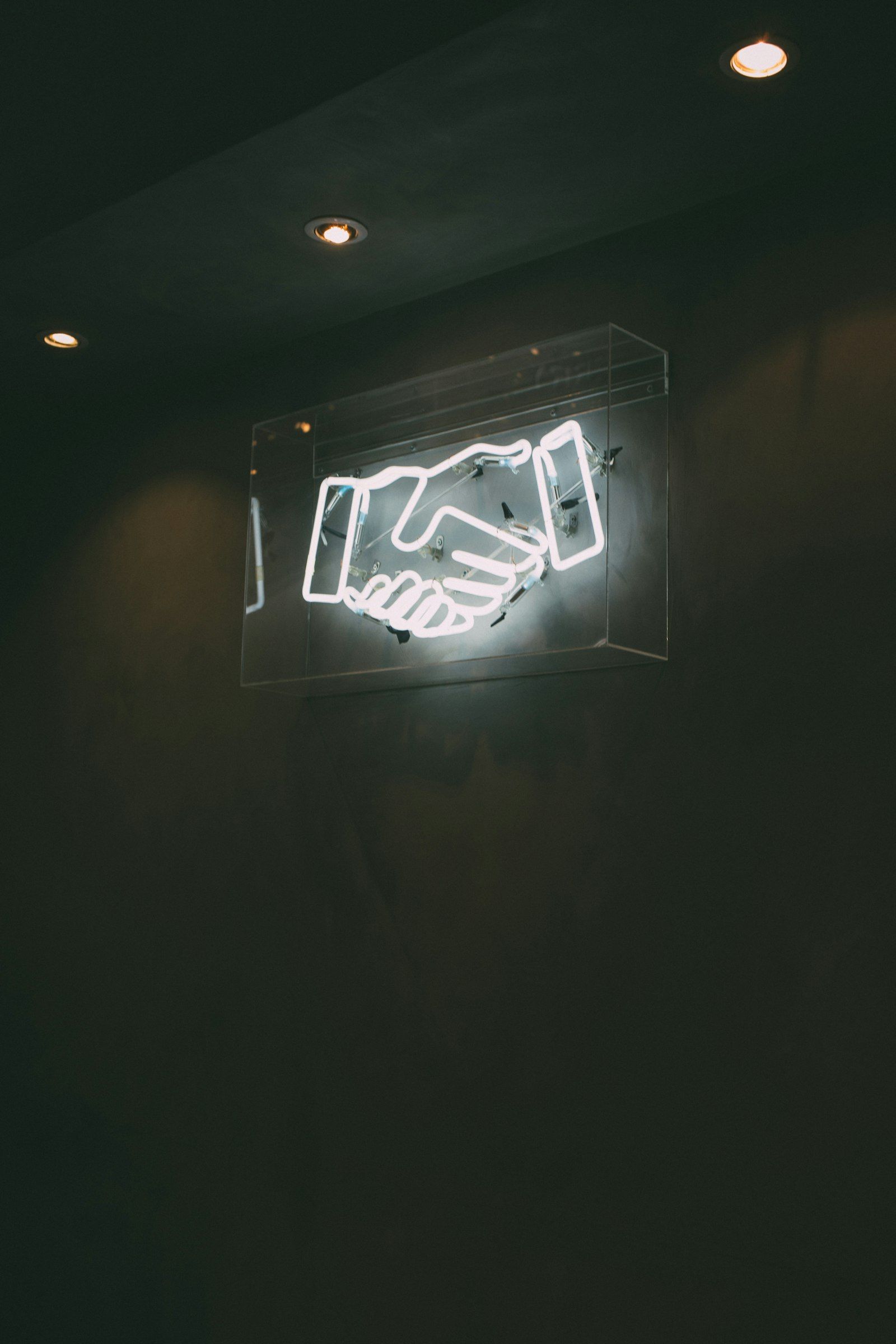 An illuminated neon sign depicting two hands shaking, symbolizing agreement or partnership. The sign is encased in a transparent box and mounted on a dark wall, emphasizing its visibility.
