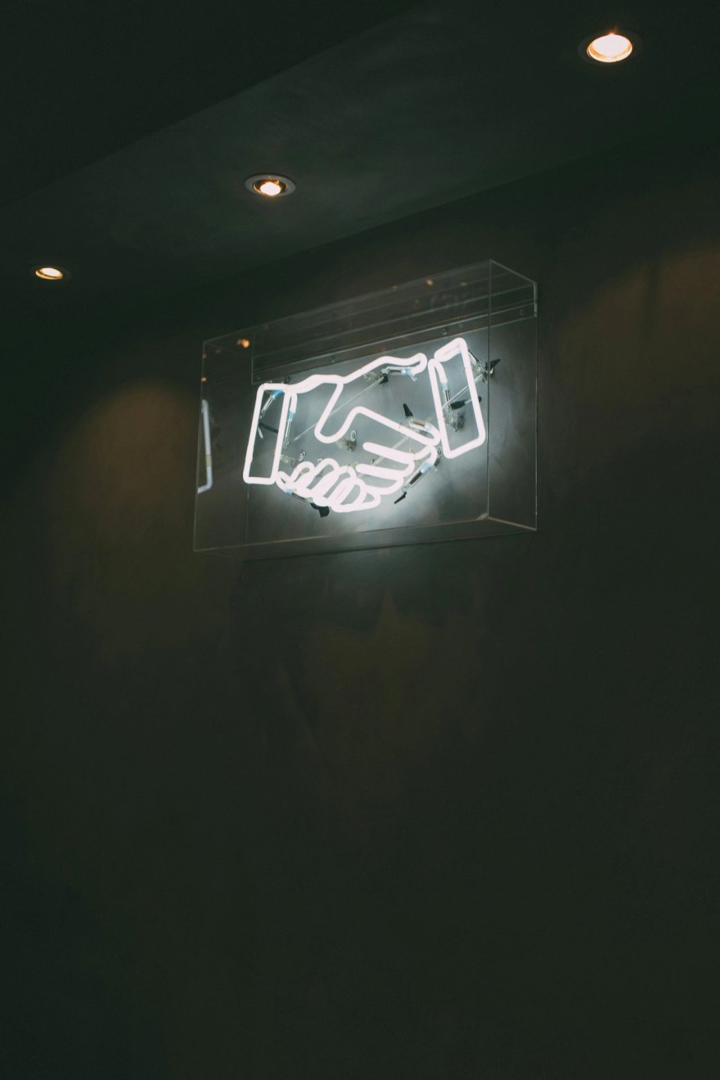 An illuminated neon sign depicting two hands shaking, symbolizing agreement or partnership. The sign is encased in a transparent box and mounted on a dark wall, emphasizing its visibility. — Credit: Photo by charlesdeluvio / Unsplash