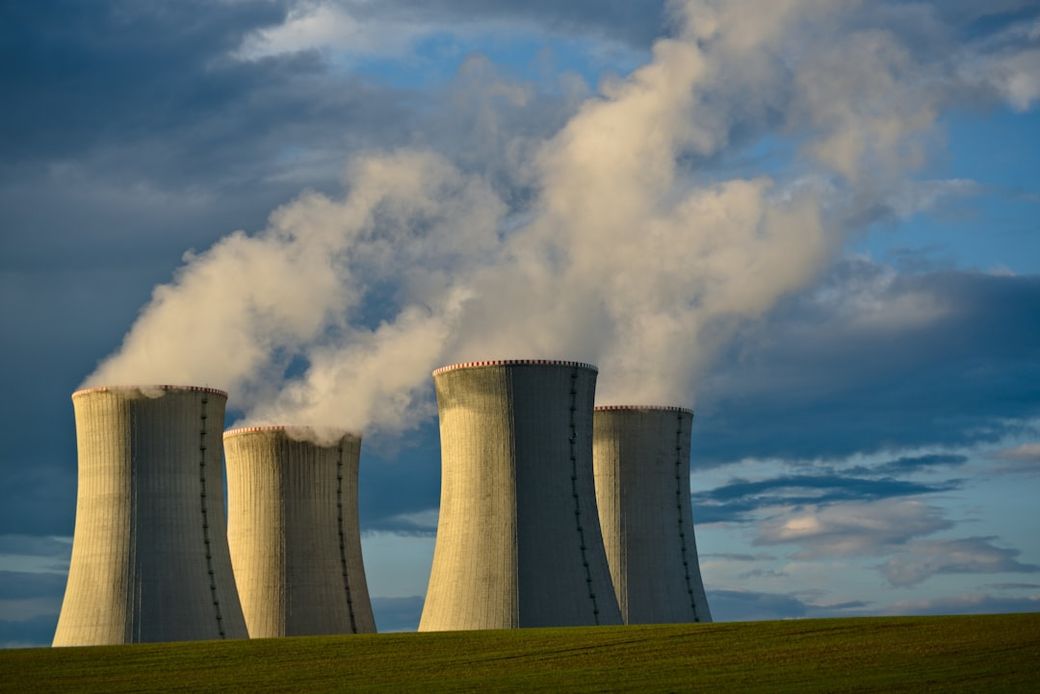 The image depicts four cooling towers of a nuclear power plant, releasing steam into the atmosphere against a backdrop of blue skies and scattered clouds. The landscape features a green field in the foreground, emphasizing the industrial aspect of the energy generation process. — Credit: Photo by Lukáš Lehotský on Unsplash