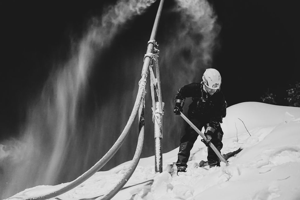 A lone worker is seen operating a snow-making machine at night, with a backdrop of snow-covered terrain. The scene captures the efforts involved in maintaining ski slopes during winter. — Credit: Photo by Jimmy Nilsson Masth on Unsplash