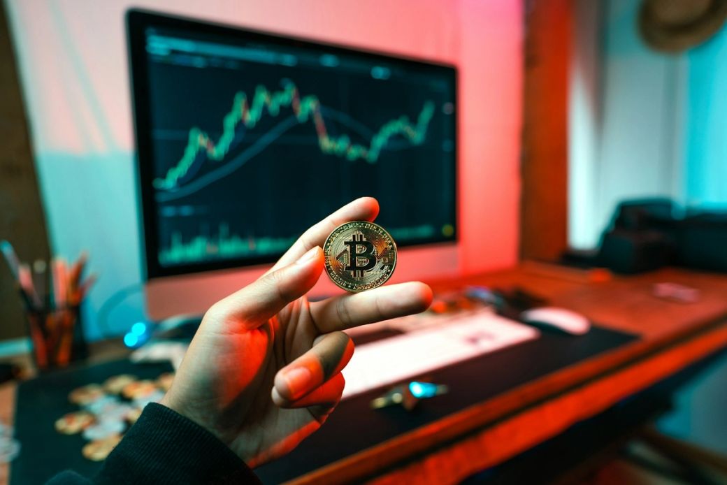 The image shows a person holding a Bitcoin in the foreground, with a trading chart displayed on a computer monitor in the background. The setting has an ambient, colorful lighting that emphasizes the focus on cryptocurrency trading. — Credit: Photo by Art Rachen / Unsplash