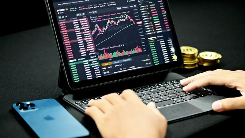 The image shows a close-up of a person using a tablet to display a cryptocurrency trading platform, with various charts and data visible on the screen. A mobile phone and gold coins are positioned nearby, indicating an active trading environment.