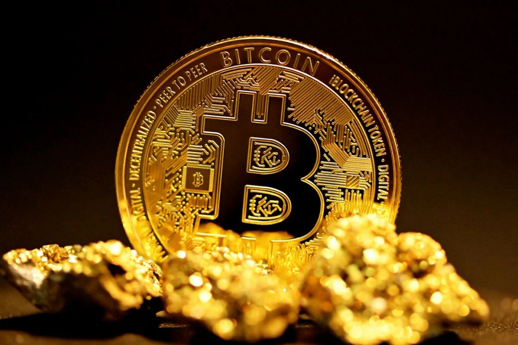 An image depicting a prominent Bitcoin cryptocurrency coin placed atop a cluster of gold nuggets. The vibrant gold and detailed design of the Bitcoin coin stands out against the dark background, symbolizing the intersection of digital and physical assets. — Credit: Photo by Kanchanara / Unsplash