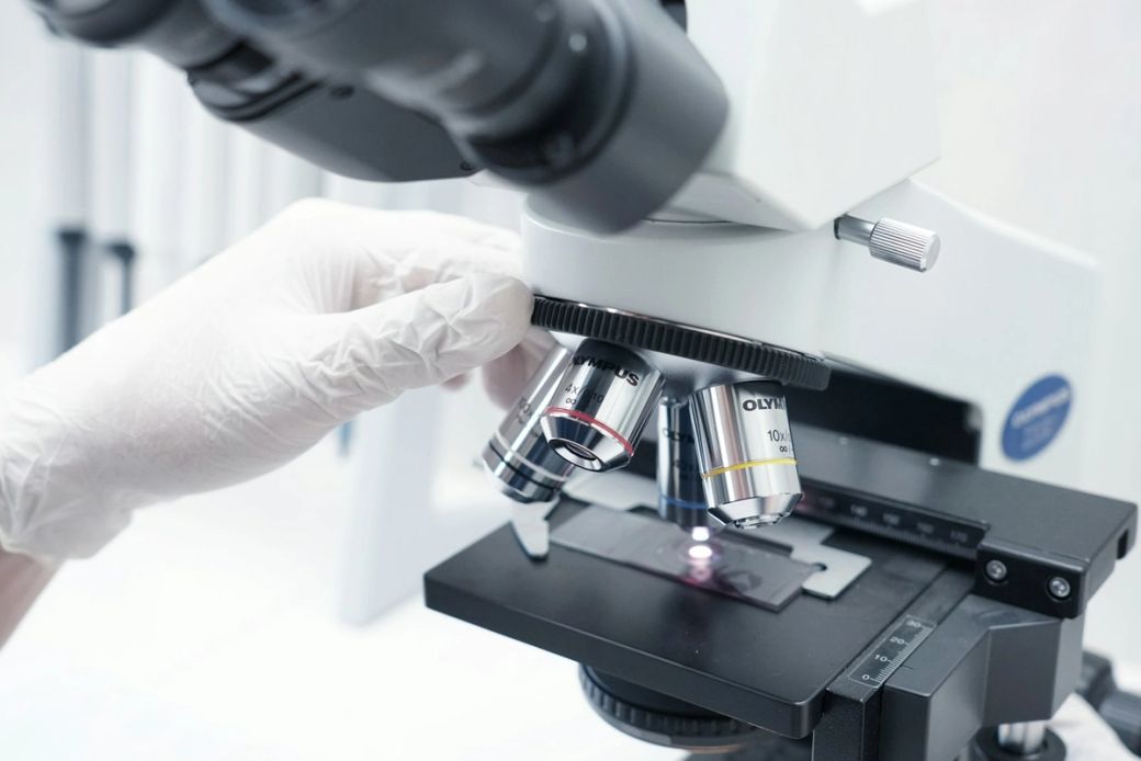 A close-up view of a laboratory microscope with a person's gloved hand adjusting the objective lenses. A glass slide is positioned on the stage, highlighting a scientific examination in progress. — Credit: Photo by Indra Projects / Unsplash
