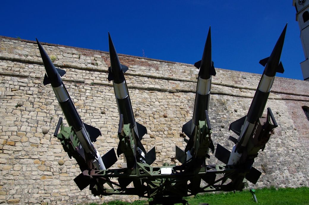 The image depicts a military missile system positioned in front of a stone wall, showcasing multiple missiles mounted on a launcher. The clear blue sky contrasts with the military hardware, emphasizing its imposing structure. — Credit: Photo by Lothar Boris Piltz on Unsplash