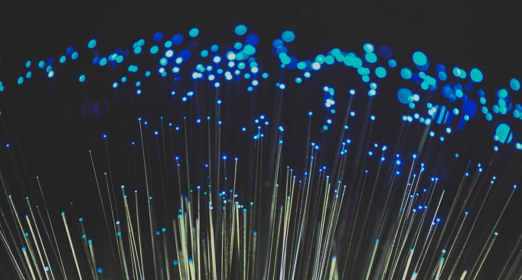 The image showcases a close-up view of fiber optic cables illuminated with blue light, conveying a sense of speed and technology. The focus on the fiber strands highlights their role in modern telecommunications. — Credit: Photo by Compare Fibre / Unsplash