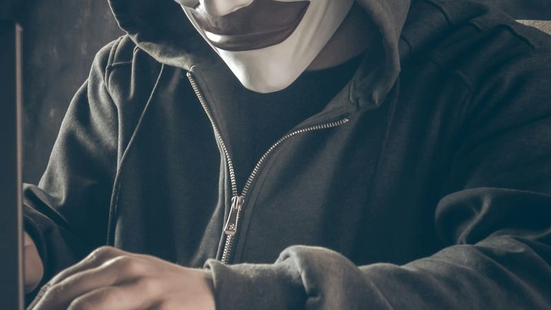 The image depicts a person wearing a hoodie and a mask, sitting in front of a laptop. The figure appears to be focused on the screen, evoking a sense of secrecy or anonymity.