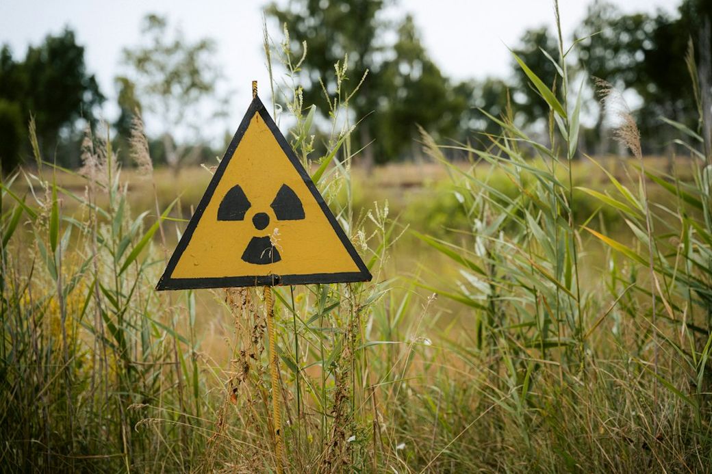 A cautionary sign indicating the presence of radioactive materials, prominently displayed in a grassy area surrounded by trees. The yellow triangular sign features a recognizable radiation symbol. — Credit: Photo by Kilian Karger on Unsplash