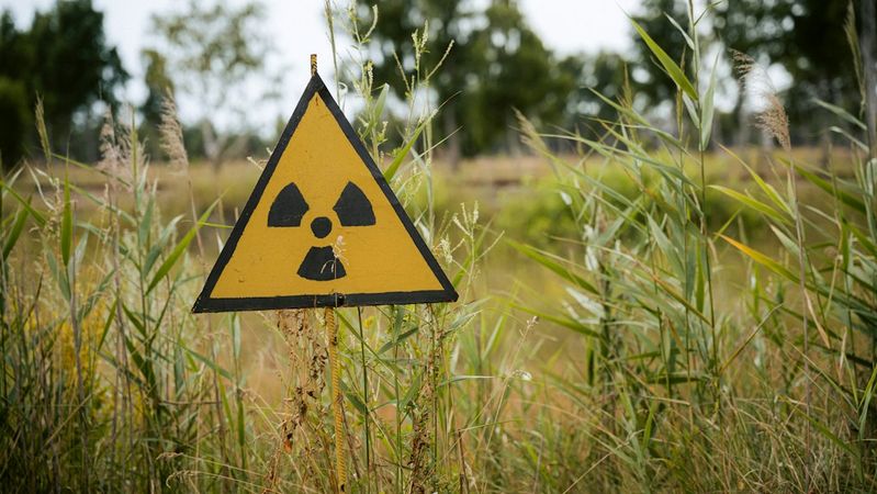 A cautionary sign indicating the presence of radioactive materials, prominently displayed in a grassy area surrounded by trees. The yellow triangular sign features a recognizable radiation symbol.