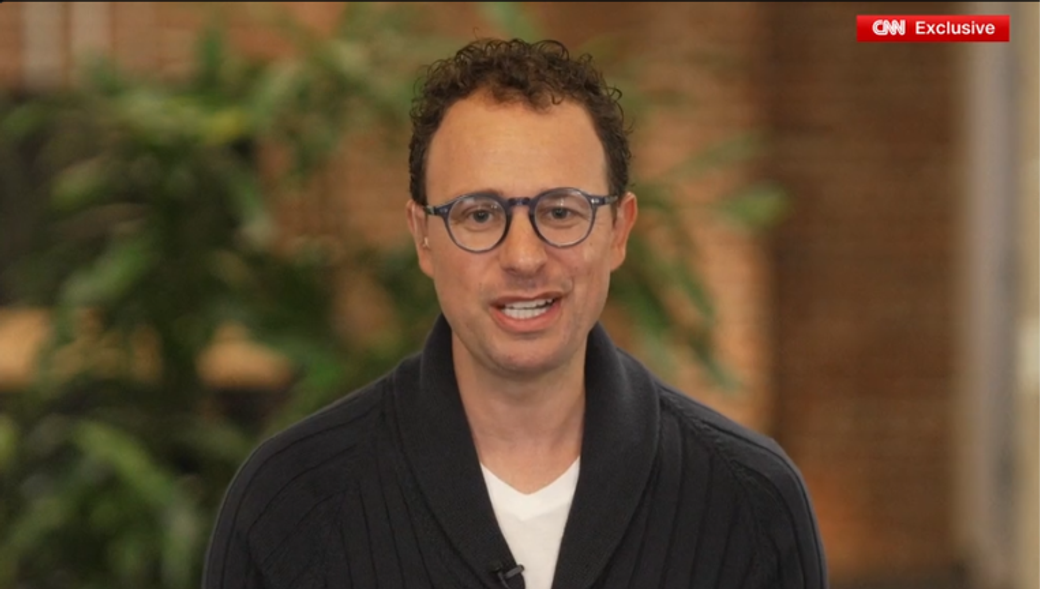 The image shows a man with curly hair and glasses speaking directly to the camera in an indoor setting with greenery in the background. He appears confident and is wearing a navy sweater over a white shirt, suggesting a casual yet professional demeanor.