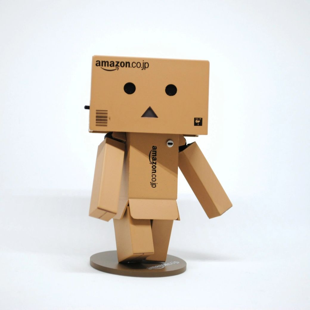 The image features a stylized cardboard robot figure with a boxy head, resembling a character often associated with Amazon. The robot has simple facial features and is positioned against a plain white background. — Credit: Photo by Nik / Unsplash