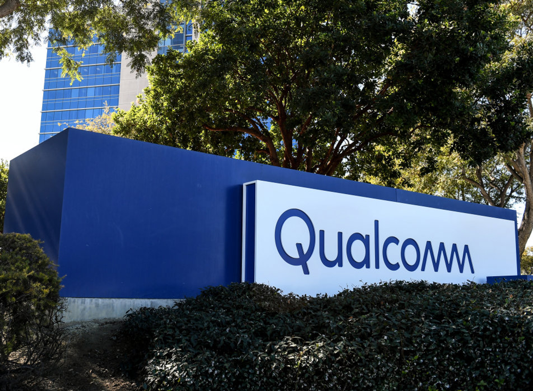 The image features the sign of Qualcomm, prominently displayed in front of a modern building surrounded by trees and shrubs. The sign, which is blue and white, indicates the presence of the technology company in a corporate setting.