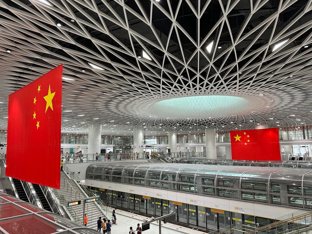 The image shows the interior of a modern transportation hub characterized by its intricate architectural design. Large Chinese flags are prominently displayed, emphasizing the national identity within the space. — Credit: Photo by Zhe ZHANG on Unsplash