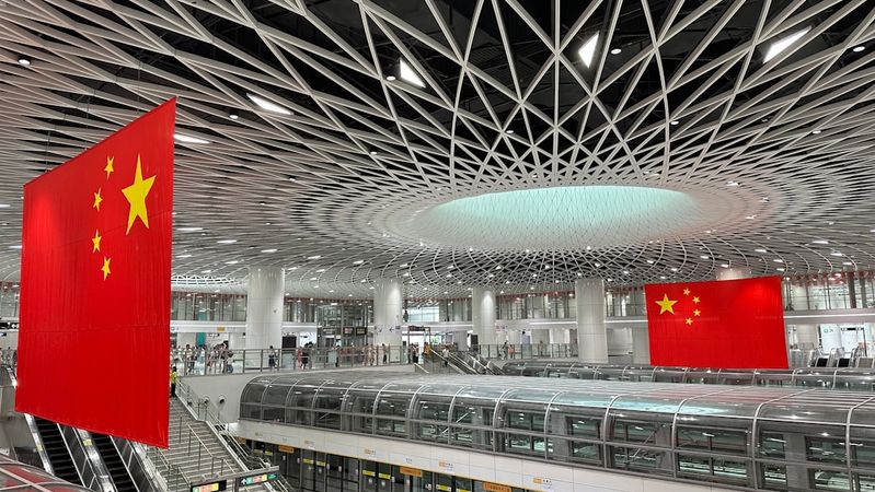 The image shows the interior of a modern transportation hub characterized by its intricate architectural design. Large Chinese flags are prominently displayed, emphasizing the national identity within the space.