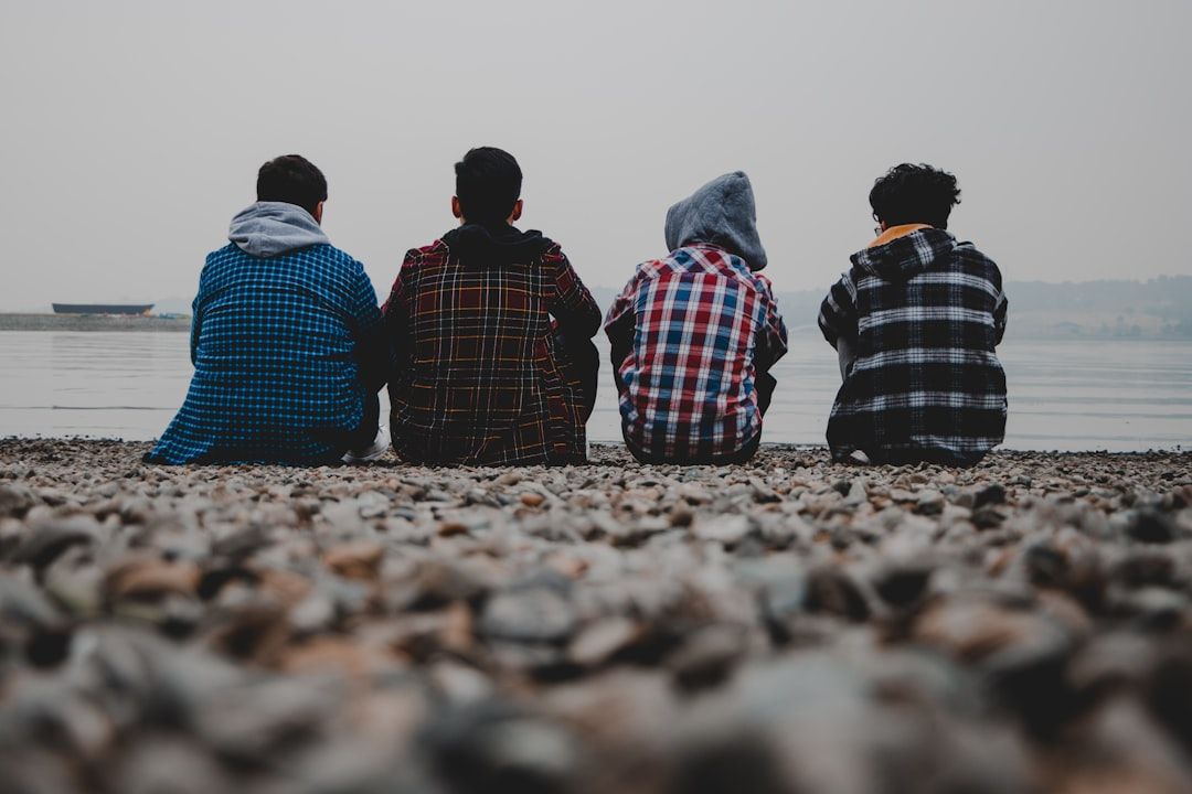The image depicts four individuals sitting on a rocky riverbank, facing the water. Their silhouettes are visible against a misty backdrop, creating a serene and contemplative atmosphere.