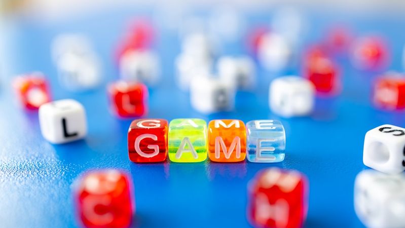 The image features colorful plastic game pieces arranged on a blue surface. The central pieces spell out the word 'GAME' in a playful manner, surrounded by other scattered letters.