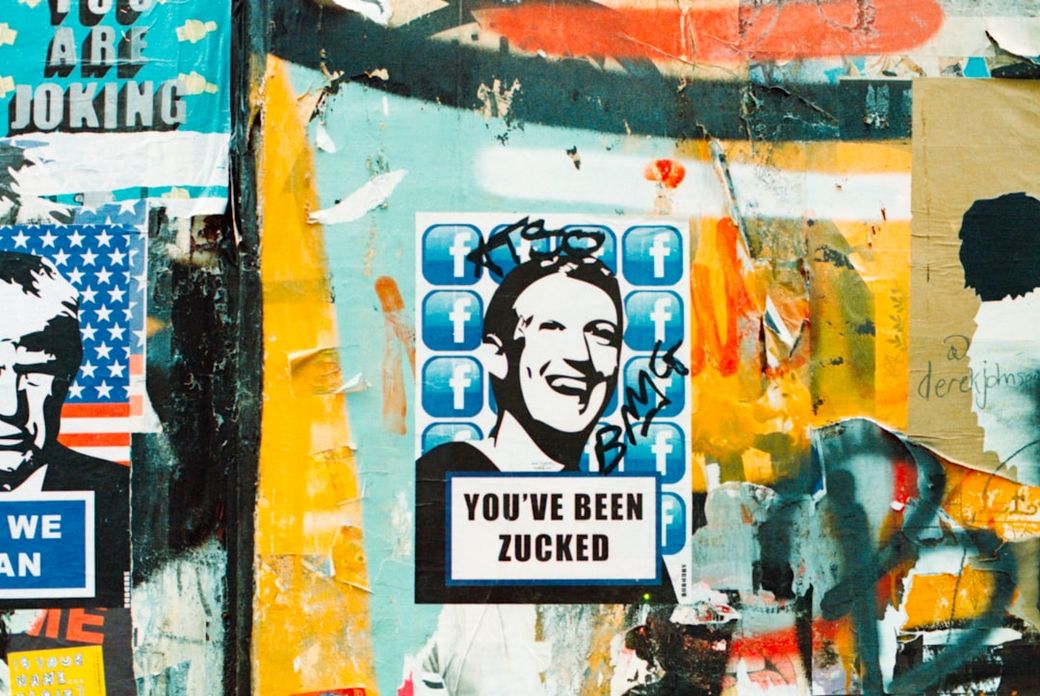 The image depicts a graffiti-covered wall featuring a prominent sticker of a smiling man resembling Mark Zuckerberg, with the text 'YOU'VE BEEN ZUCKED' prominently displayed. The background is a collage of various other posters and graffiti, creating a vibrant urban art scene. — Credit: Photo by Annie Spratt on Unsplash