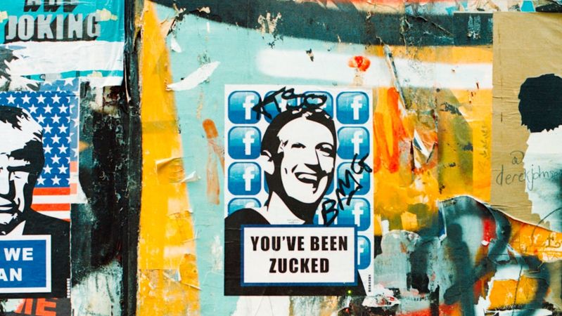 The image depicts a graffiti-covered wall featuring a prominent sticker of a smiling man resembling Mark Zuckerberg, with the text 'YOU'VE BEEN ZUCKED' prominently displayed. The background is a collage of various other posters and graffiti, creating a vibrant urban art scene.