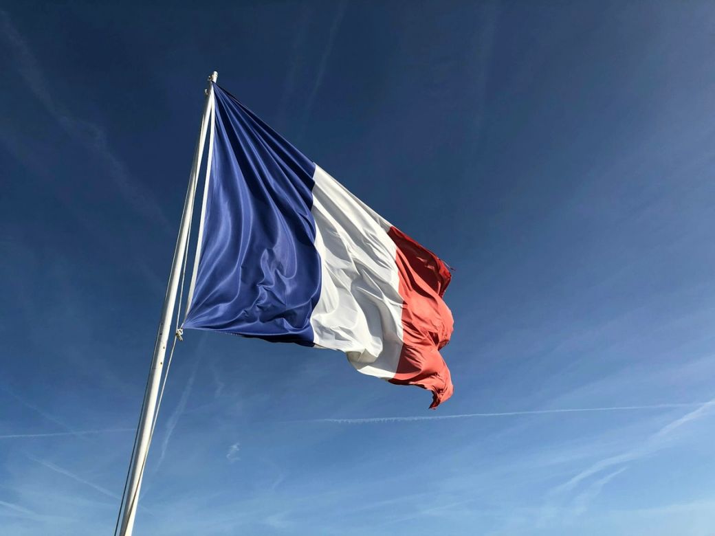 The image depicts the national flag of France, prominently waving against a clear blue sky. The tricolor flag features vertical bands of blue, white, and red. — Credit: Photo by Anthony Choren / Unsplash