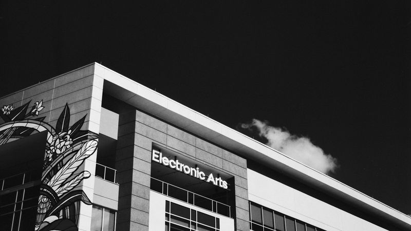 The image features the exterior of the Electronic Arts building, showcasing a modern architectural design with a stylized mural on the side. Clear skies with a single cloud can be seen above.