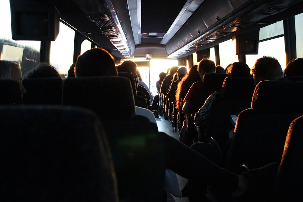 The image shows the interior of a bus, filled with passengers seated in rows, as sunlight streams in from the front. The warm light creates a cozy atmosphere, highlighting the faces of people commuting. — Credit: Photo by Ash Gerlach on Unsplash